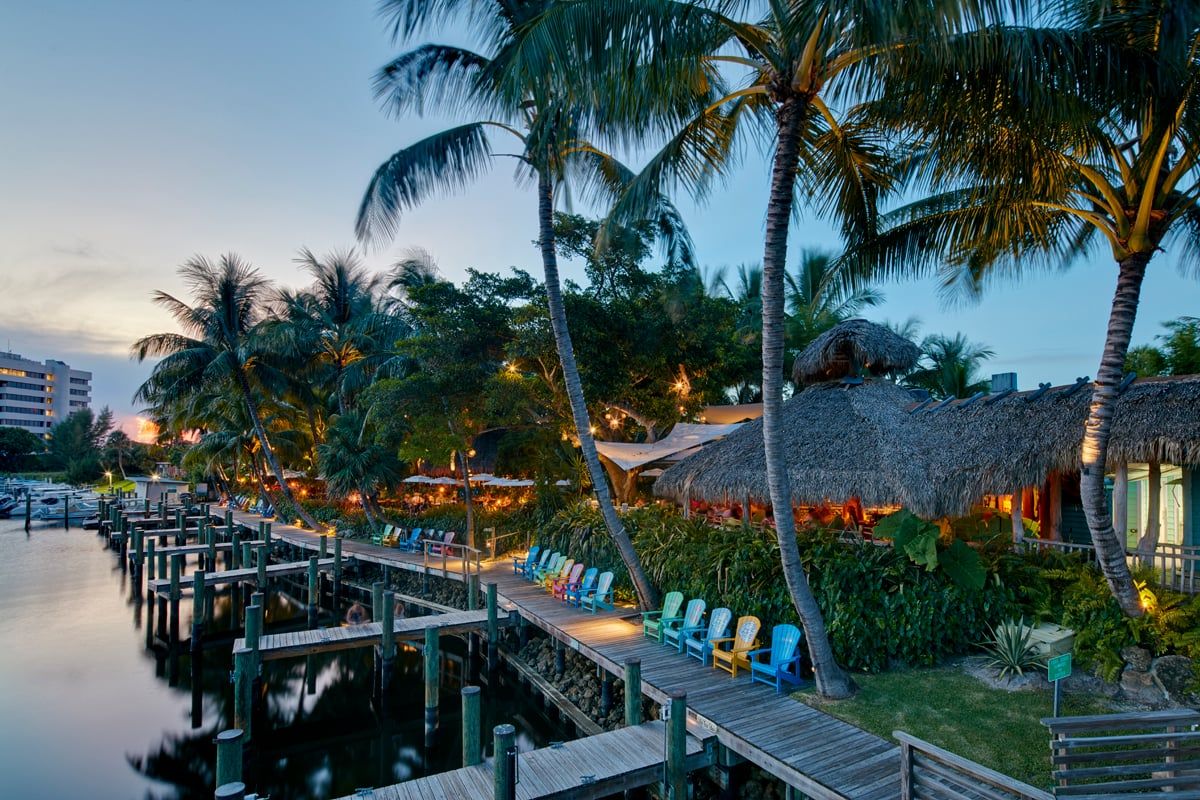 Wide blue-hour view of the whole venue from across the water — boardwalk, rainbow chairs, thatched tiki roofs, string-lit dining