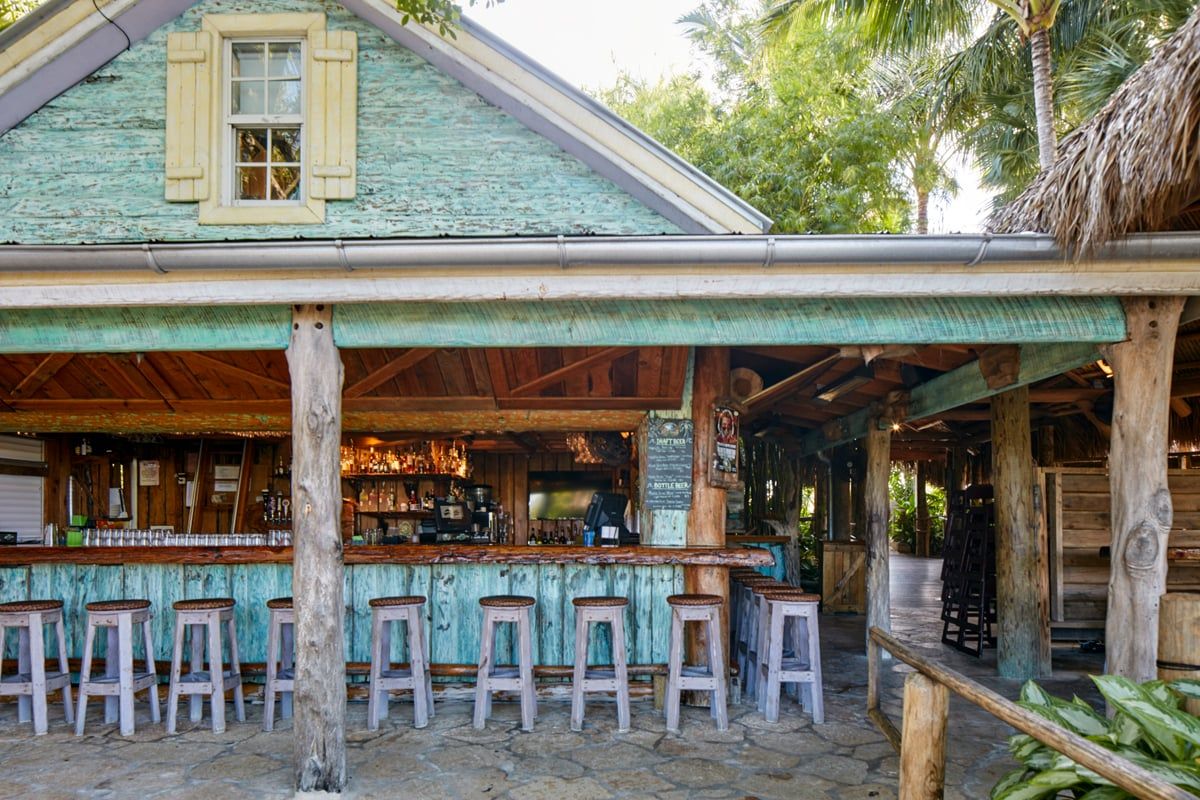 The weathered green-clapboard coquina bar building with a long row of rustic wooden barstools
