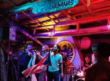 A band performing in the tiki room at night under the Guanabanas sign and surfer mural, with a guest waving a flag in the crowd