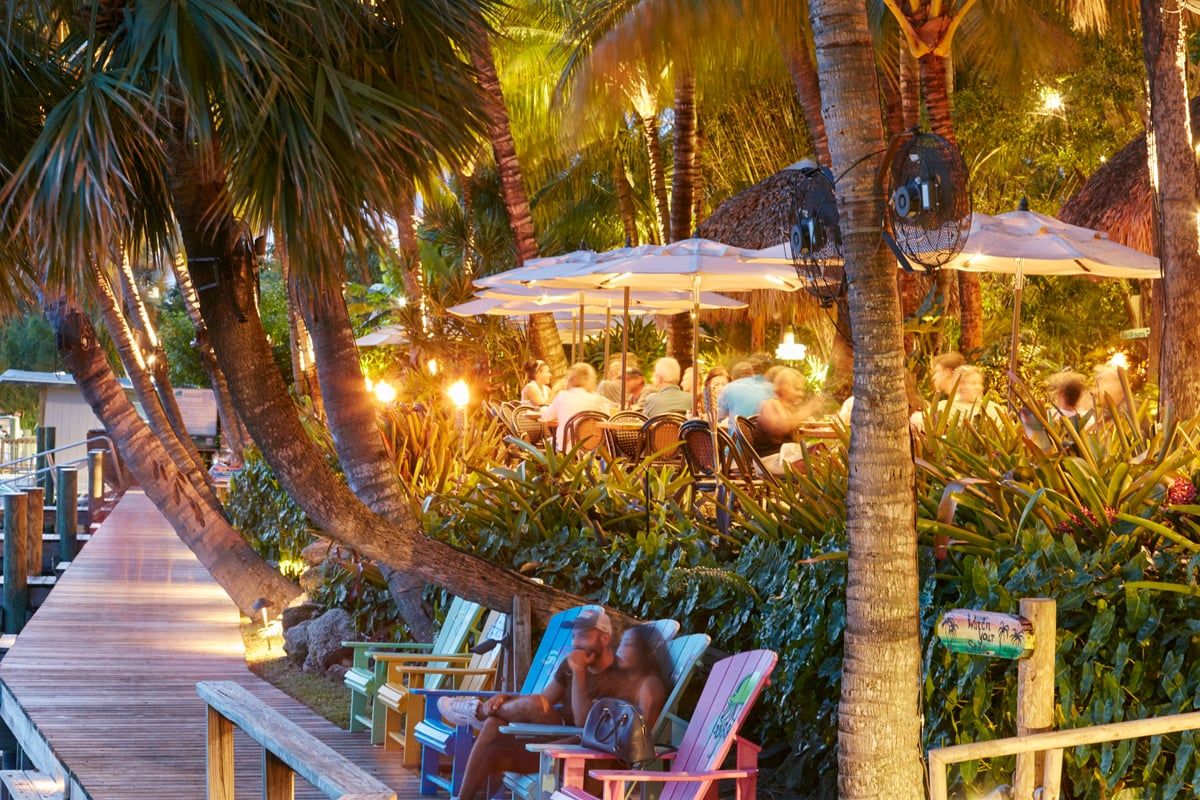 The string-lit open-air patio at dusk — guests dining under white market umbrellas among palms and lush foliage, warm bistro lights overhead