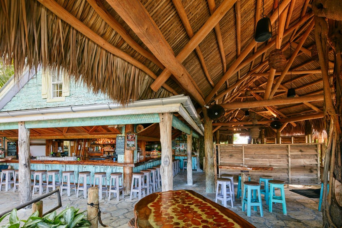 Inside the thatched tiki bar looking up at the hand-built palm-frond roof, with a turquoise-washed bar and a pizza on a live-edge wood table